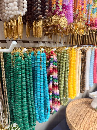 A display of colorful beaded necklaces and bracelets hanging on a rack, featuring vibrant green, blue, pink, yellow, and white beads alongside gold-toned jewelry.