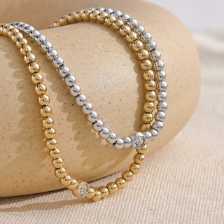 Two beaded bracelets, one gold and one silver, on a beige stone surface.