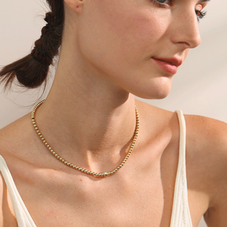 A close-up lifestyle shot of a woman wearing a delicate gold beaded necklace, showing the jewelry on her neck against a soft, warm neutral background.