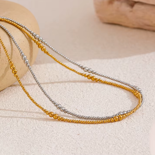Two beaded chains, one gold and one silver, arranged on a textured beige surface with a neutral stone prop.