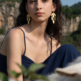 A woman wearing large gold leaf-shaped dangle earrings, photographed outdoors against a blurred rocky landscape background.
