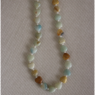 A beaded necklace made of polished, irregular tumbled stones in light blue, green, beige, and brown, separated by small white spacer beads, displayed on a textured beige fabric background.