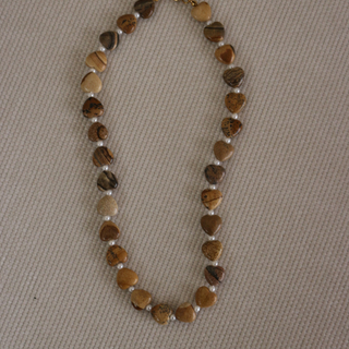 A beaded necklace made of polished brown and tan stones with small white spacer beads, laid flat on a textured beige fabric background.