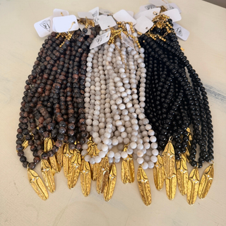 Three strands of beaded prayer beads with gold arrow-shaped pendants, displayed side by side on a light beige surface. The left strand features dark brown beads, the middle strand features off-white/cream beads, and the right strand features black beads.