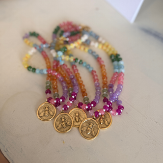 A set of multicolor beaded necklaces with gold coin charms, arranged on a light beige surface.