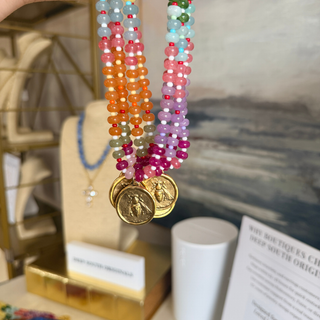 A close-up of a beaded necklace with multiple strands of colorful round beads in orange, pink, purple, blue, green, and white, hanging from gold-toned coins on a display stand.