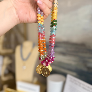 A hand holds up a pair of multi-strand beaded necklaces with gold-toned coin charms. The necklaces feature colorful beads in yellow, orange, red, pink, purple, green, and blue, with the gold coins showing detailed engravings.