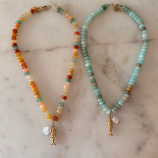 Two beaded fish necklaces displayed on a light marble surface. The necklace on the left features a multicolor bead strand with gold-toned fish pendants.