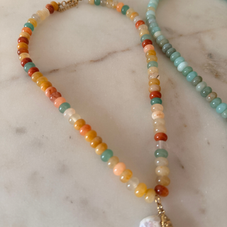 A beaded necklace made of small round gemstone-like beads in warm orange, yellow, peach, green, and cream tones, with a gold clasp and a white pearl-like pendant, displayed on a light marble surface.