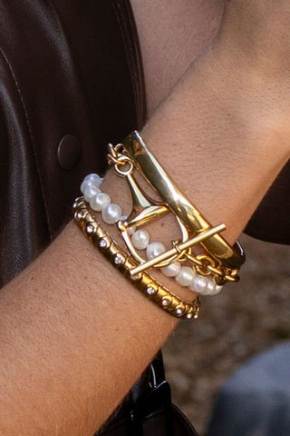 A close-up of a person's wrist wearing a stack of gold and pearl bracelets, including a gold chain bracelet, a pearl beaded bracelet, and a gold bracelet with clear rhinestones.