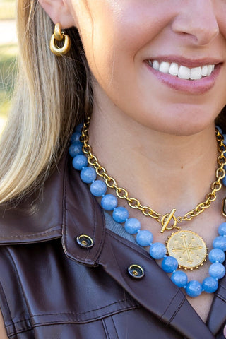 A close-up lifestyle shot of a woman wearing a layered necklace with light blue beads and gold chain, along with a gold hoop earring and a brown leather jacket.