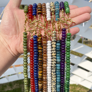 A hand holds up multiple beaded bracelets with gold toggle clasp chains. The bracelets feature various colorful stone beads including green, red, blue, brown, white, black-spotted, and purple, shown against a blurred outdoor background.