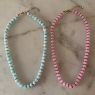 Two beaded necklaces displayed on a light marble surface. One necklace is made of light blue and white round beads with a gold clasp, and the other is made of pink and white round beads with a gold clasp.