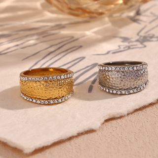 Two textured band rings displayed on a beige fabric surface. One ring is gold with clear rhinestones, and the other is silver with clear rhinestones.