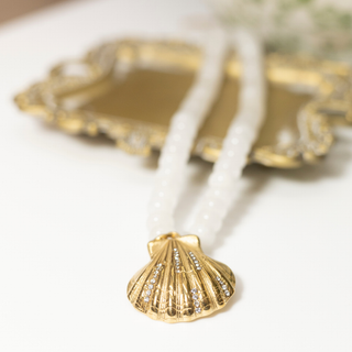 A gold shell-shaped pendant necklace with small clear rhinestones, hanging from a strand of white beads, with an ornate gold tray blurred in the background.