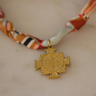 Terra Benedict Necklace with a cream, orange and rust patterned fabric cord and ornate gold-tone cross medallion pendant.